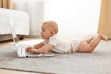 Side view portrait of curious baby lying on floor and looking at opened laptop computer display with interest, studying modern technology, posing against window in light room.