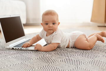 Indoor side view portrait of cute child lying on floor near laptop with blank screen and looking at camera, wearing white t shirt, posing in living room against window.