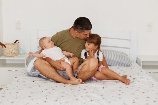 Indoor Shot Of Happy Father Wearing Casual Style T Shirt Sitting On Bed With Crossed Legs With His Kids, Kissing Her Elder Daughter While Holding Infant Baby In Hands.