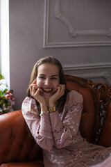 Fototapeta na wymiar Young beautiful woman in pink dress and stylish accessories sits on brown leather sofa on white wall background. Portrait of happy smiling girl with red lips resting at home and looking at camera. 