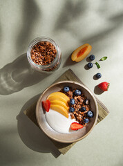 Delicious natural yogurt with homemade granola, peach, strawberry and blueberry in a bowl and jar with muesli on a green background morning shadows. Healthy and nutritious breakfast. Top view