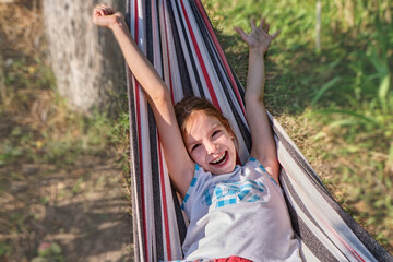 A happy little girl lies in a hammock in the garden in the summer. A child in a hammock during the holidays smiles in the park against the background of green grass. High quality photo