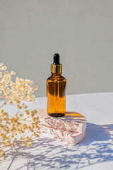 Amber glass dropper bottle with metallic lid on a podium with dry plants on light background....