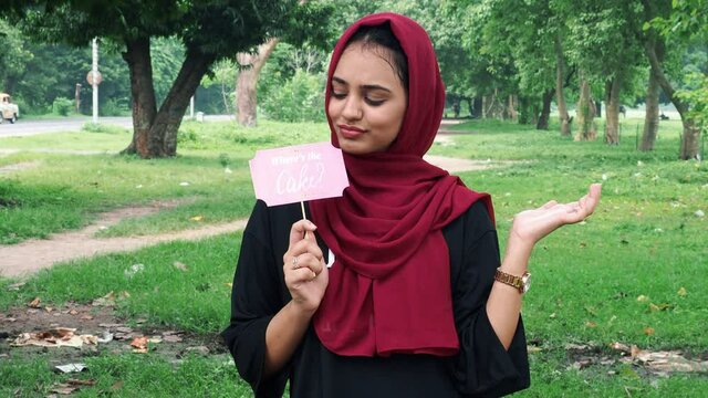 Young Afghan women posing in hijab using pink photo booth props written where is the cake.Young happy woman holds a paper on a stick and smile.