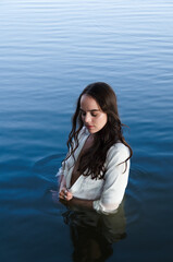 Thoughtful and calm young woman in white shirt is standing in water, looking away.