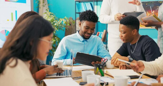 Manager in blue shirt supervises co-workers, does not apply himself to duties, surfs the Internet on a tablet in the office, shows a colleague funny videos, tells a story, distracts him from work