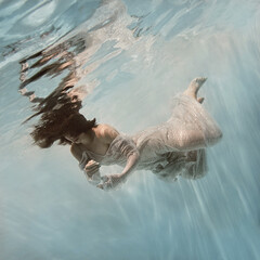 A girl in a blue dress swims underwater on a light background as if in weightlessness