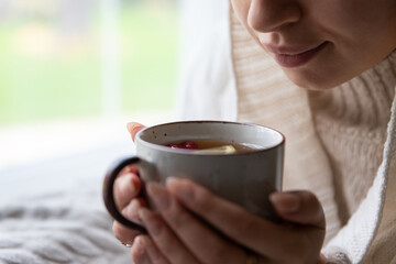 Young woman drinking hot tea while resting at home in morning, dreaming sitting by window covered with warm blanket. Autumn time concept. Girl with sweater relaxing while drinking purifying herbal tea
