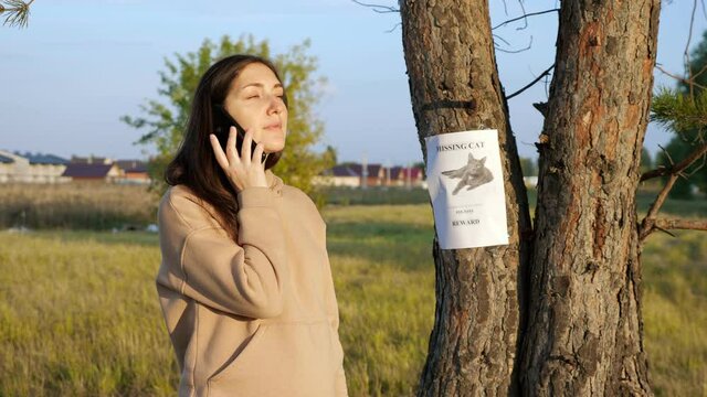 Smiling woman in hoodie dials number from missing cat poster hanging on tree trunk to inform owners of found pet in autumn close view