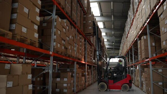 Warehouse loader standing at rack, lifting goods in boxes up and setting high on empty space. Storehouse machinery during work, forklift driver placing huge pallet of cardboard boxes on rack