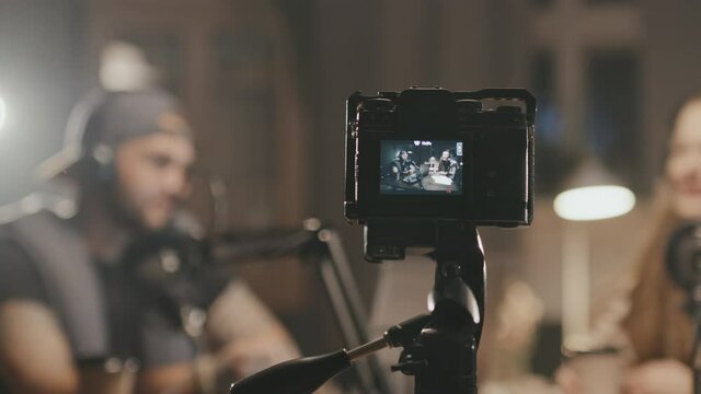 Tracking shot of video camera filming people in headphones talking into microphones while recording their podcast or show