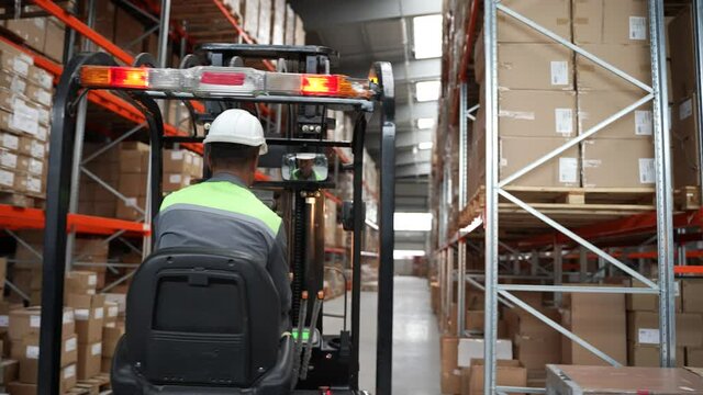 Back view of forklift driver riding between racks in storehouse with pallet of cardboard boxes to place on shelf. Male employee in helmet driving warehouse loader during work