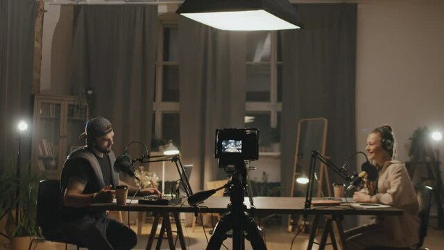 Dolly-in shot of young man and woman sitting at table in living room lit by studio lights and recording podcast or online show