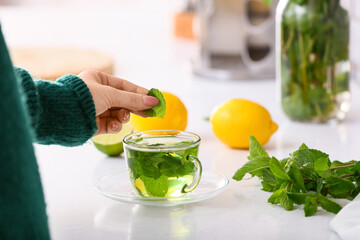 Woman putting mint leaf into glass cup with tea in kitchen, closeup