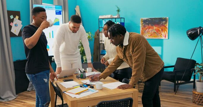 Four hard-working employees talk about the statistics created, man sips coffee, conclude that they have finished preparing analysis, happy and high-five each other for their joint work