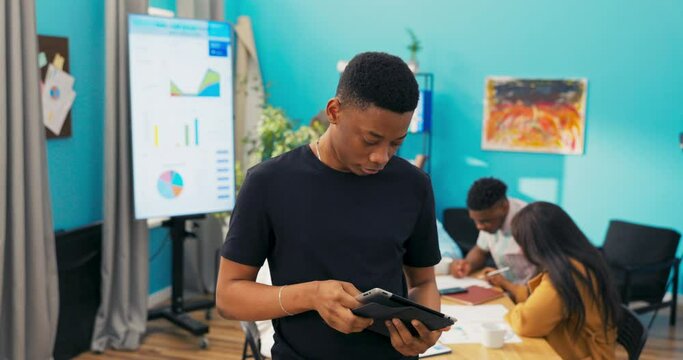 Proud, happy and smiling, dark-skinned erasmus student is group leader, supervising project work of colleagues in corporate office. The boy browses tablet raises head crosses arms on chest