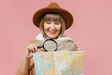 Close up traveler tourist mature elderly senior lady woman 55 years old wear brown shirt hat scarf hold examine map with magnifying glass isolated plain pastel light pink background studio portrait