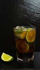 Cuba Libre cocktail with brown rum, lemon juice, coke and ice cubes, decorated with fresh lime slice on modern black stone tray