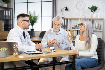 Caucasian medical worker white lab coat and eyewear prescribing treatment to mature couple that sitting at his cabinet. Aged man and woman visiting family doctor for health examination.