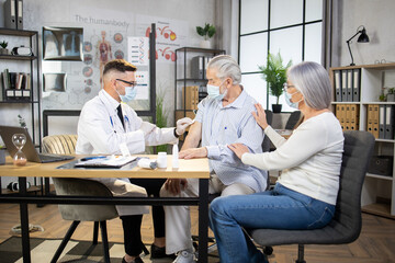 Caucasian male doctor in face mask and gloves doing vaccination for senior man at office. Mature wife cheering her husband during health treatment at clinic.