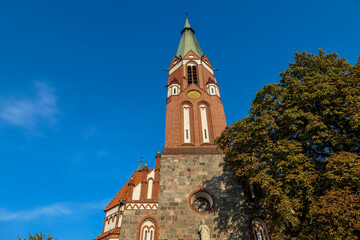 Fototapeta na wymiar A close up of a bell tower of a church in Sopot, Poland. The church is surrounded by high, lush trees. The sky above is blue and cloudless. Spirituality.