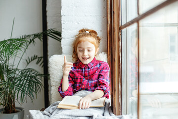 Little girl child reads an interesting book. Child on the windowsill. Big window. Warm blanket.