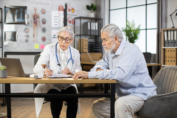 Confident female mature physician writing in clipboard while sitting at desk and taking anamnesis from aged male patient, complaining to health condition during regular checkup at clinic.