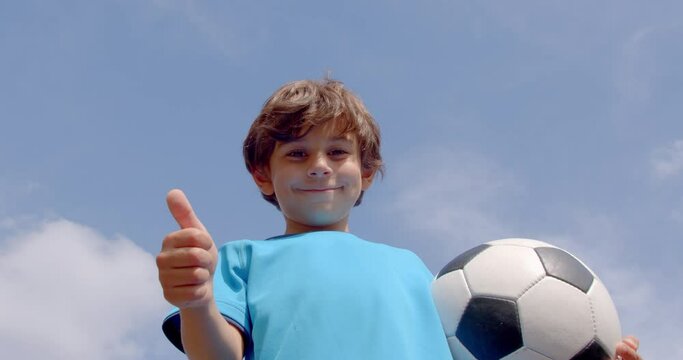 A cute 7-8 years old boy in football equipment and a ball in hand showing thumb up on the blue sky, 4k slow motion low angle view