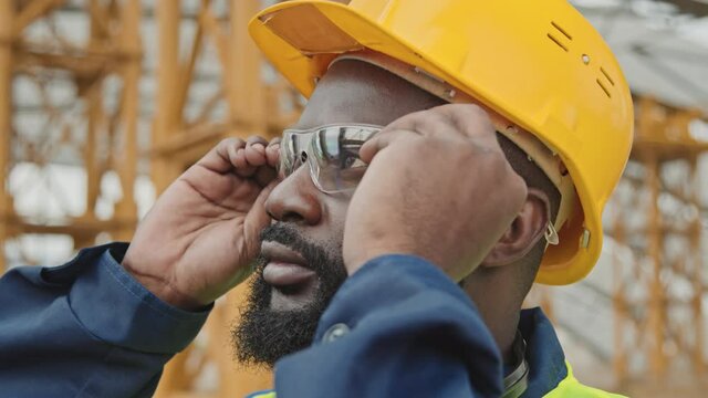 Close up shot of African-American male worker in hard hat putting on safety goggles at construction site