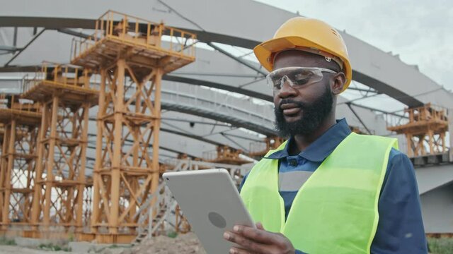 Tracking shot of busy African-American male construction site supervisor in safety vest, hard hat and goggles using tablet outside. Unfinished building with scaffolding in background