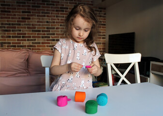 A little girl with brown eyes and wavy hair at a large white table sculpts from pink plasticine. Plasticine in pink, orange, blue and green colors is on the table. Classes in the garden and at home. 