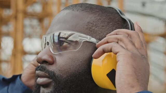 Extreme close up of black male worker in safety goggles putting on noise-canceling earmuffs at construction site