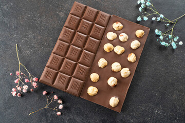 Milk chocolate bar with whole hazelnut kernels, flowers on black background. Healthy food concept, delicious chocolate sweets for kids, diet, sugar