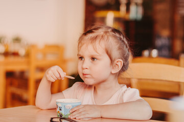 Little girl sitting at the table alone , holding spoon and eating ice cream.