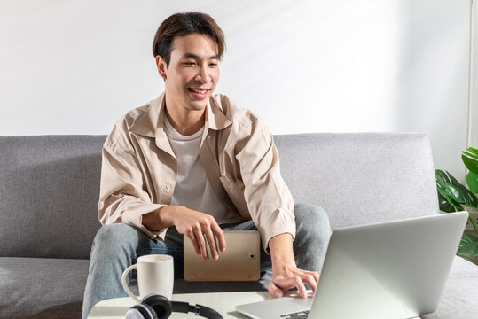 A young happy Handsome Asian man as a designer sitting on a couch at a home apartment looking at laptop and tablet cellphone screens for online shopping, online social searching, or video conference