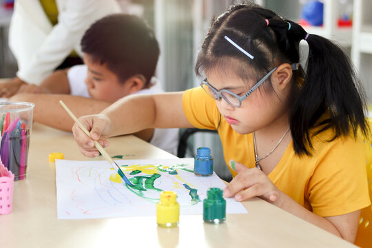 Happy children having fun with friends during study at school, girl with down syndrome concentrate painting on paper in art classroom, education of kids with physical disability and intellectual conce