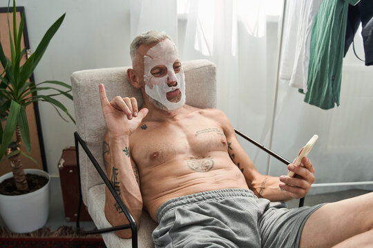 Man with facial mask sitting at the armchair and making selfie at the smartphone