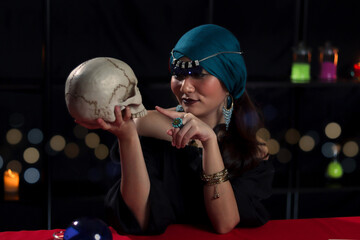 Dark witch casting a spell on skull, mysterious magnificent beautiful woman in black dress holding...