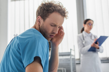 curly patient suffering from headache near blurred doctor in hospital