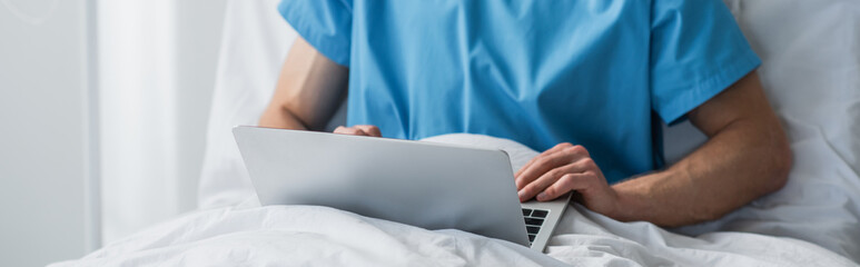 cropped view of freelancer in patient gown using laptop in hospital bed, banner