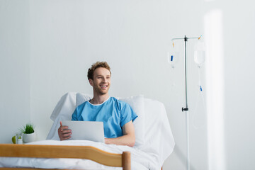 cheerful freelancer in patient gown using laptop in hospital bed