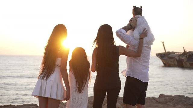 Relaxed multigenerational family admiring golden sunset on Mediterranean sea shore talking pointing at sunken old ship. Back view of happy Caucasian mother father and daughters in sunshine outdoors
