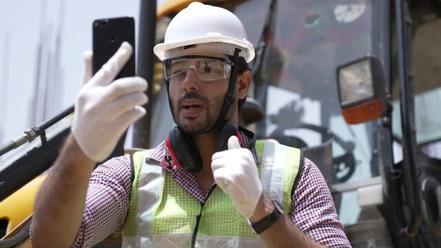 Smart Indian engineer holding smartphone and making video call with costumer about building information,Asian man standing in work site,he feeling happy and smile, video call and internet technology.