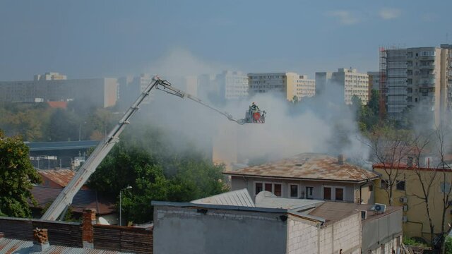 Team of firefighters on service to put out fire from burning house in urban landscape. Firemen using platform truck and water extinguisher helping with flames and smoke from building