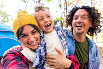 young happy family portrait camping outdoor with tent in autumn nature. mom, dad and little child making a pov selfie into the wild. people having fun together hugging each other smiling. lifestyle - obrazy, fototapety, plakaty