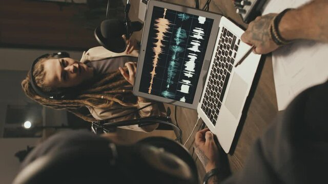 Handheld vertical shot of young man and woman sitting at table at home or studio and discussing topic on their podcast