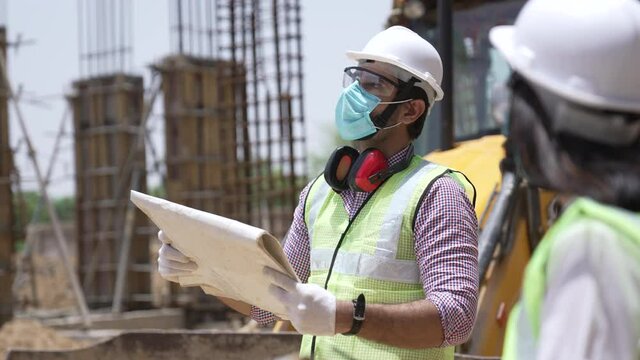 Young Indian man engineer matching details with blue prints and site plans on site.Asian male wearing protective eyeglass with helmet making plans for their project on construction site outdoor.
