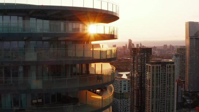 Ascending shot of tall modern round apartment building with rings of terraces around. Revealing cityscape against setting sun. London, UK
