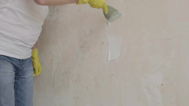 Plasterer man holding putty knife is spackling with putty plaster patching a hole in white wall. worker's hand in yellow gloves holding putty knife patching aligning wall. Home renovation concept