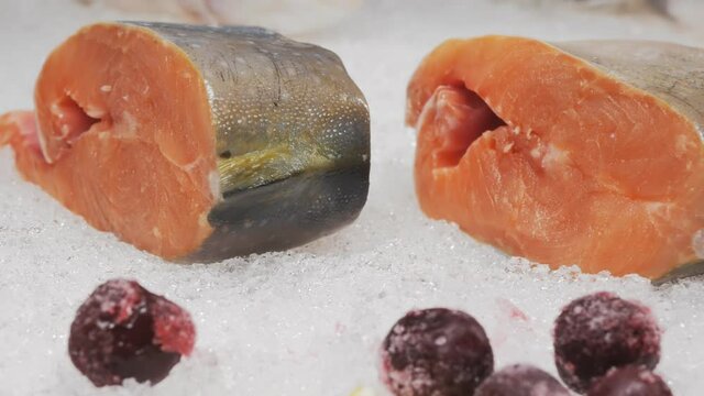 Close-up, dolly shot, DOF: pieces of salmon fresh on the frozen frozen snow lie in a seafood market showcase. In the background, lemon, berries and greens